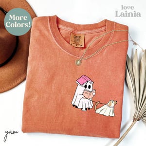 Op de afbeelding: Koraalrood Comfort Colors T-shirt met een cartoongeest die een boek leest en een hond uitlaat, beide in spookkostuums. Het shirt heeft de tekst "More Colors!" en "love Lainia". Een gouden ketting is over het shirt gedrapeerd.