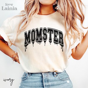 May include: An ivory short-sleeve t-shirt featuring the black text MOMSTER in a collegiate font with a dripping effect. The shirt has a crew neckline and is paired with blue denim jeans. The text love Lainia is visible in the top left corner.