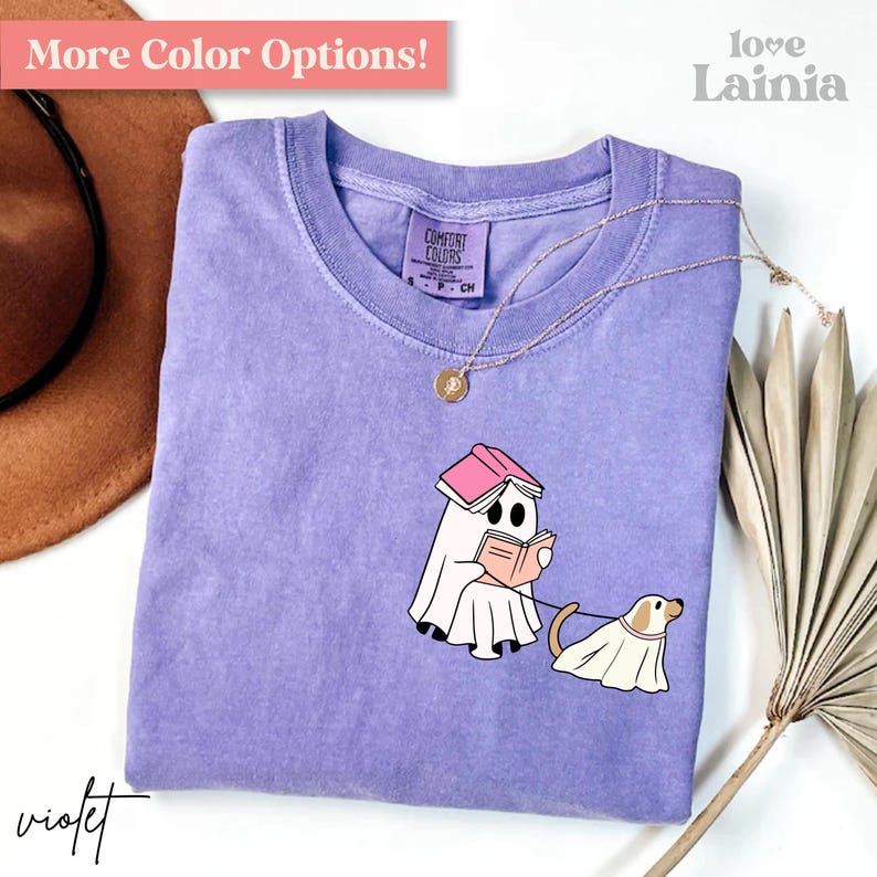 Op de afbeelding: Violet Comfort Colors T-shirt met een cartoongeest die een boek leest en een spookhond uitlaat. De shirt heeft een ronde hals en korte mouwen. De tekst "More Color Options!" staat bovenaan. Een gouden ketting ligt op het shirt.