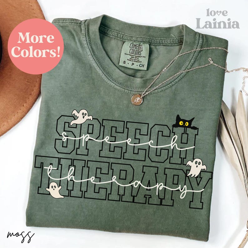 May include: A moss green t-shirt with the words "SPEECH THERAPY" in a black, block letter design, with a white cursive font overlay. The shirt features a small black cat and two ghost graphics. A silver necklace with a pendant is draped on the shirt.
