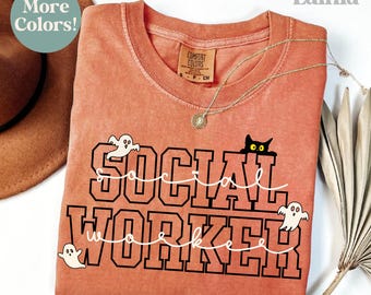 May include: A coral-colored t-shirt with the words "SOCIAL WORKER" in a black, block font, and white cursive font. The shirt features a black cat graphic and three ghost graphics. A gold necklace is draped across the shirt.