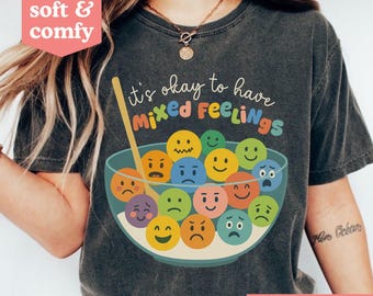 Könnte beinhalten: Dunkelgraues T-Shirt mit dem Text "it's okay to have mixed feelings" über einer Schüssel mit bunten, ausdrucksstarken Gesichtern. Das Shirt hat auch die Worte "soft & comfy" in einem rosa herzförmigen Kasten und "MORE COLORS!" in einem roten Kasten.