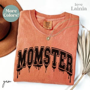 May include: A terracotta-coloured short-sleeve t-shirt with a crew neckline. The shirt features the word MOMSTER printed in a large, black, varsity-style font with a dripping effect at the bottom. The inner collar tag reads Comfort Colors.