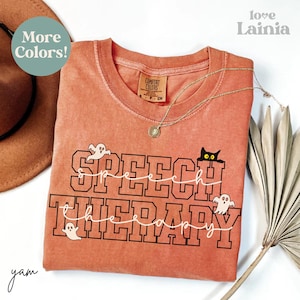 May include: An orange Comfort Colors t-shirt with the words "SPEECH THERAPY" in a black collegiate font, with a white cursive font overlay. The shirt features a black cat and ghost graphics. A gold necklace and a hat are also visible.