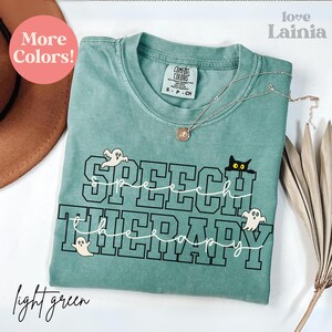 May include: A light green t-shirt with the words "SPEECH THERAPY" in a black and white design, featuring ghost and cat graphics. The shirt has a crew neck and a small tag. A silver necklace with a pendant is draped on the shirt.