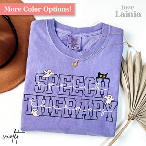 May include: A lilac Comfort Colors t-shirt featuring the text "SPEECH THERAPY" in black, with ghost and cat illustrations. The shirt has a crew neck and is adorned with a gold necklace. The top left corner reads "More Color Options!" in pink.