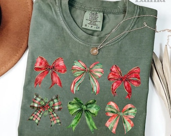 May include: A moss green t-shirt featuring six watercolor bows in red, green, and plaid patterns. The shirt has a Comfort Colors tag and a delicate silver necklace. The words "love Lainia" and "moss" are also visible.