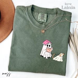 Op de afbeelding: Mosgroen T-shirt met een afbeelding van een geest die een boek leest en een hond uitlaat, beide in spookkostuums. Het shirt heeft een Comfort Colors-label en een ketting. De woorden "love Lainia" staan rechtsboven.