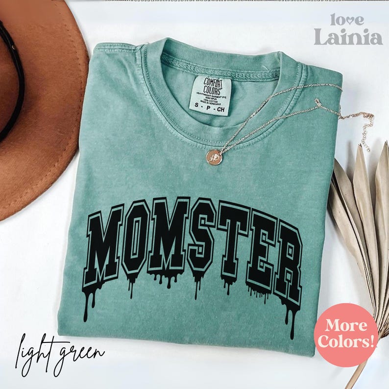 May include: A sage green short-sleeve t-shirt with a crew neckline and a Comfort Colors label. The shirt features the black text MOMSTER in a varsity-style font with a dripping effect. A thin silver-tone chain necklace with a small pendant rests on top.