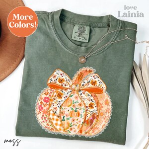 May include: A moss green Comfort Colors t-shirt featuring a large pumpkin design with a floral bow. The pumpkin is decorated with orange, white, and pink floral patterns. The shirt has the text "More Colors!" in an orange circle.