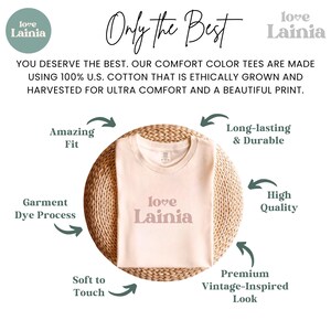 May include: A cream-coloured t-shirt with the text "love Lainia" in a soft pink hue. The shirt is displayed on a woven circular surface. Text around the shirt highlights features like amazing fit, soft touch, and a vintage-inspired look.
