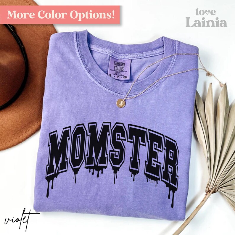 May include: A violet short-sleeved t-shirt with a crew neckline featuring the word MOMSTER in black collegiate-style lettering with a dripping effect. The shirt has a Comfort Colors label at the inner neck and is displayed with a gold-toned necklace.