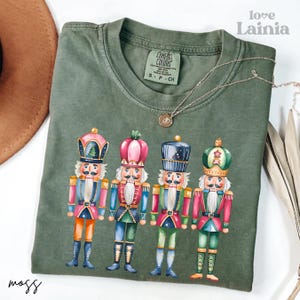 May include: A moss green Comfort Colors t-shirt featuring a colorful graphic of four nutcrackers. Each nutcracker is dressed in a different uniform with pink, blue, green, and gold accents. The words "love Lainia" are in the top right corner.