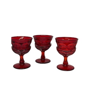 May include: Three ruby red wine glasses with a textured, geometric pattern. Each glass has a rounded bowl, a slender stem, and a circular base. The glasses are arranged in a row against a white background.