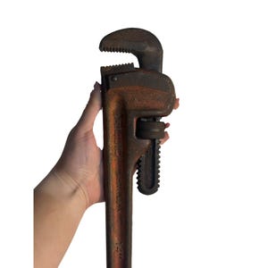 May include: A large, rusty orange and black pipe wrench. The wrench has a toothed jaw and a textured handle. The tool is held in a hand against a white background. The wrench is a heavy-duty tool.