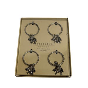 May include: A set of four Pottery Barn Seaside Napkin Rings. Each ring is silver-toned and features a charm with a palm tree and dolphin design. The rings are displayed in a cardboard box with the text "OUTLET Seaside Napkin Rings Set of four".