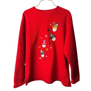 May include: A red long-sleeve fleece sweatshirt with an embroidered design of snowmen wearing hats and scarves. The snowmen are playing musical instruments and are surrounded by snowflakes and stars. The sweatshirt is hanging on a metal hanger.