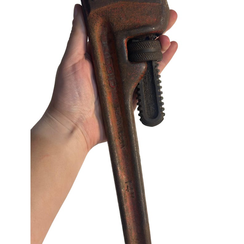 May include: A rusty, orange and black pipe wrench. The wrench is held in a hand and has a textured handle and adjustable jaws. The wrench is a Ridgid brand and is a 3.8 cm model.