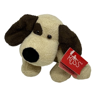 May include: A plush dog toy with cream-colored fur, dark brown ears, and a black nose. The toy features dark brown patches around the eyes and a red tag attached to the side that reads Russ Tannenbaum with a small butterfly icon and heart patterns.