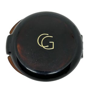 May include: A round, dark brown tortoiseshell compact case with a glossy finish. The center of the lid features a gold-colored logo consisting of the overlapping letters C and G. The compact has a smooth, circular design with visible side hinges.