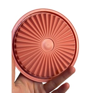 May include: A coral-colored, round Tupperware lid with a sunburst design. The lid is held in a hand, showcasing its textured, ridged surface. The center features a raised, circular detail.