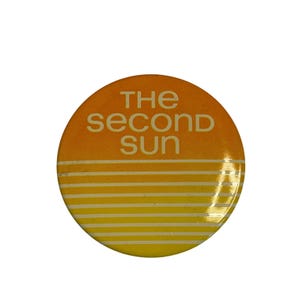 May include: A round button with a gradient design of orange and yellow, featuring the text "THE SECOND SUN" in white. The button has horizontal stripes at the bottom, creating a sunset effect.