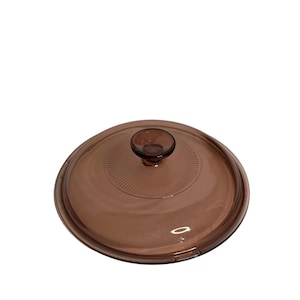 May include: A brown glass lid with a circular design and a small, round handle. The lid is transparent, allowing a view of the contents below. The lid is designed for cookware.