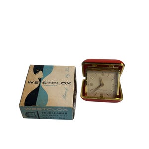 May include: Vintage Westclox Touralarm II luminous dial travel alarm clock in a red case. The clock face is white with black numerals and hands. The original box is included, featuring the Westclox logo and product details.