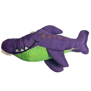 May include: A plush shark toy featuring a purple upper body with orange triangle patterns and a bright green underbelly. The shark has a stitched black mouth with white teeth, a dorsal fin, two side fins, and a tail fin, all in purple fabric.