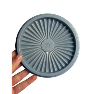May include: A round, light gray Tupperware lid with a ridged, fan-like design radiating from the center. The lid is held by a hand against a white background. The Tupperware logo is visible in the center.