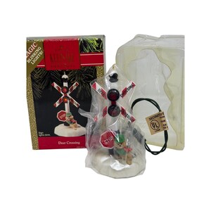 May include: A Hallmark Keepsake Christmas ornament featuring a miniature railroad crossing sign with blinking lights. The sign has red and white details, with a small deer figurine at the base. The box reads "Deer Crossing."