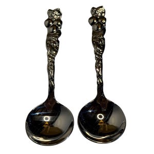 May include: Two silver-toned serving spoons. Each spoon has a round bowl and a long, ornate handle. The handle features a detailed, textured design with a figure at the top. The spoons are reflective, with a polished finish.