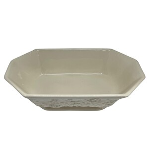 May include: An off-white ceramic bowl with an octagonal rim and a shallow basin. The exterior side features a raised, textured floral relief pattern that wraps around the base, providing a decorative element to the smooth, glossy finish.