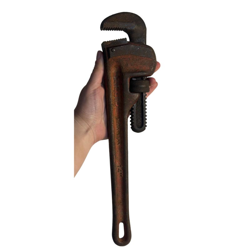 May include: A vintage 35.5 cm pipe wrench with a rusty, weathered appearance. The wrench is held in a hand, showcasing its size and design. The tool features a toothed jaw and an adjustable mechanism, typical of plumbing tools.