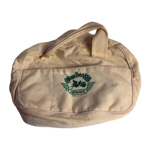 May include: A cream-colored canvas duffel bag with a top handle and a front pocket. The bag features a green embroidered logo that reads Cabbage Patch Kids AIRWAYS with a stork graphic and the text 1983 O.A.A. INC. below the logo.