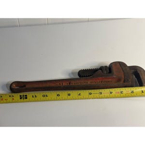 May include: A vintage red and brown pipe wrench, with a toothed jaw and adjustable mechanism. The wrench is approximately 33 cm long, with visible text on the handle. A yellow measuring tape is placed next to the wrench.