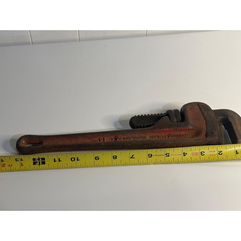 May include: A vintage red and brown pipe wrench, with a toothed jaw and adjustable mechanism. The wrench is approximately 33 cm long, with visible text on the handle. A yellow measuring tape is placed next to the wrench.