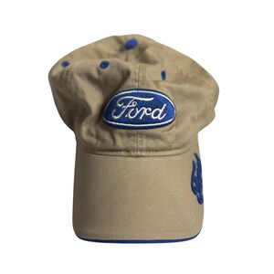 May include: A tan baseball cap featuring a blue oval patch with the white embroidered text Ford on the front. The cap includes blue eyelets on the crown, a blue trim on the edge of the brim, and additional blue embroidery on the side panel.