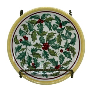 May include: A round decorative plate with a yellow rim and a white center. The plate features a pattern of green holly leaves and red berries. The plate is held by a decorative metal stand.