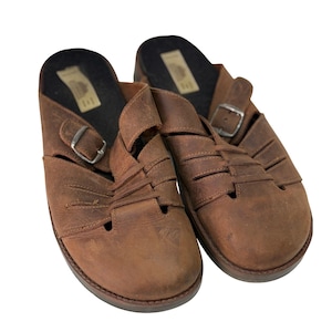 May include: A pair of brown leather clogs with a distressed finish. The clogs feature a woven design across the top, a buckle closure, and a black insole. The shoes have a rounded toe and a low heel.