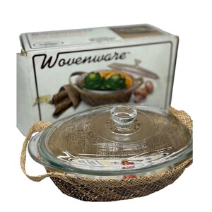 May include: An oval glass casserole dish with a clear glass lid featuring a knob handle. The dish is encased in a brown woven basket holder with two side handles. The product is displayed in front of its original Wovenware branded cardboard box.