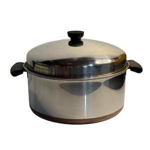 May include: A stainless steel cooking pot with a polished silver finish, featuring a matching lid with a black knob and two black side handles. The base of the pot has a copper-colored band, and the overall design is cylindrical and functional.
