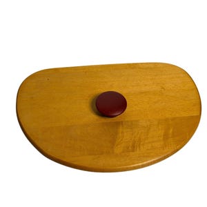 May include: A light-colored wooden cutting board with a rounded shape and a dark red, circular handle. The board has a smooth surface and a natural wood grain pattern. The handle is centered on the board.