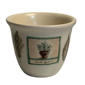 May include: Cream-colored ceramic cup with a botanical design. The cup features a rectangular image of a potted plant labeled "sage" and leaf illustrations. The cup has a slightly flared rim and a smooth, glossy finish.