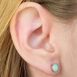 May include: A close-up of an ear with a small, oval-shaped stud earring. The earring features a pale green gemstone set in a silver or white metal setting. The earring is a simple, elegant design.