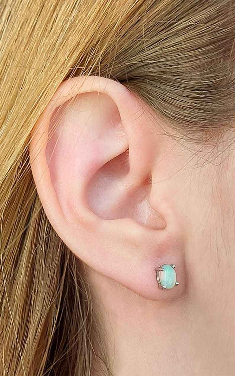 May include: A close-up of an ear with a small, oval-shaped stud earring. The earring features a pale green gemstone set in a silver or white metal setting. The earring is a simple, elegant design.