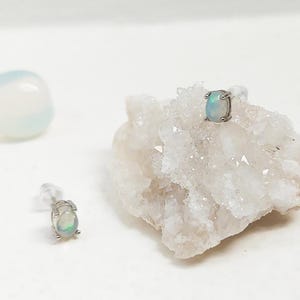 May include: Two opal stud earrings with a silver setting are displayed on a white surface. One earring is placed on a white crystal formation, and the other is positioned separately. Two white, rounded stones are also in the background.