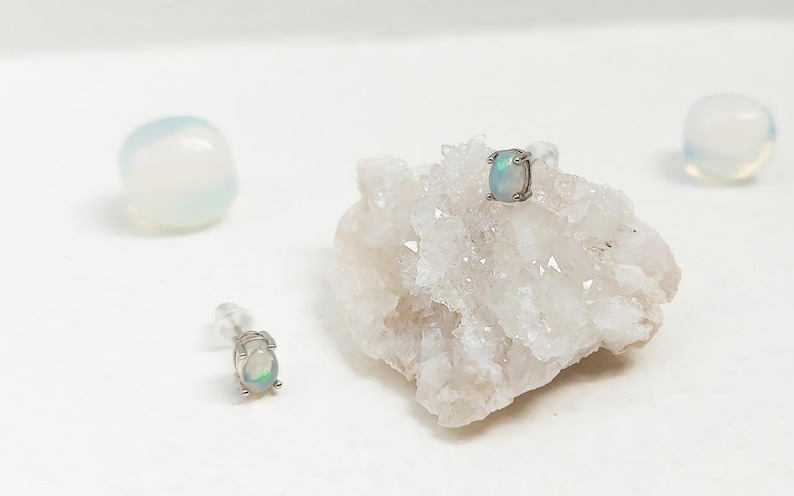 May include: Two opal stud earrings with a silver setting are displayed on a white surface. One earring is placed on a white crystal formation, and the other is positioned separately. Two white, rounded stones are also in the background.