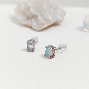 May include: A pair of oval-shaped opal stud earrings with a silver setting. The opals display a play of color, with flashes of blue, green, and pink. The earrings are set against a white background with other decorative stones.