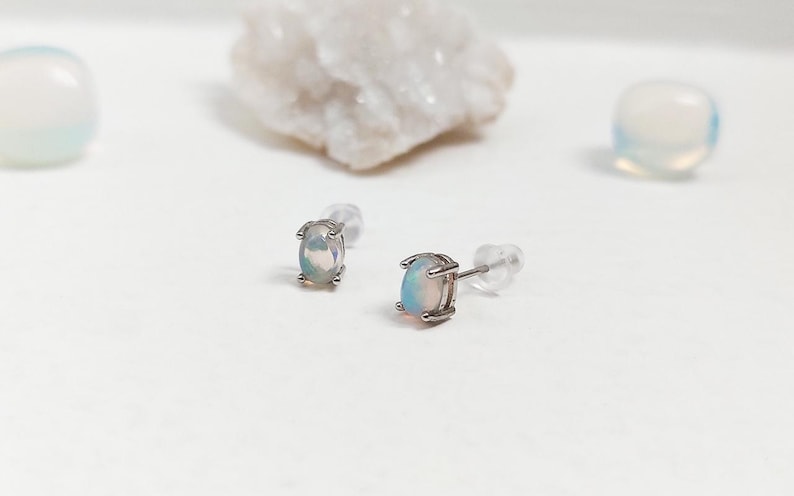 May include: A pair of oval-shaped opal stud earrings with a silver setting. The opals display a play of color, with flashes of blue, green, and pink. The earrings are set against a white background with other decorative stones.
